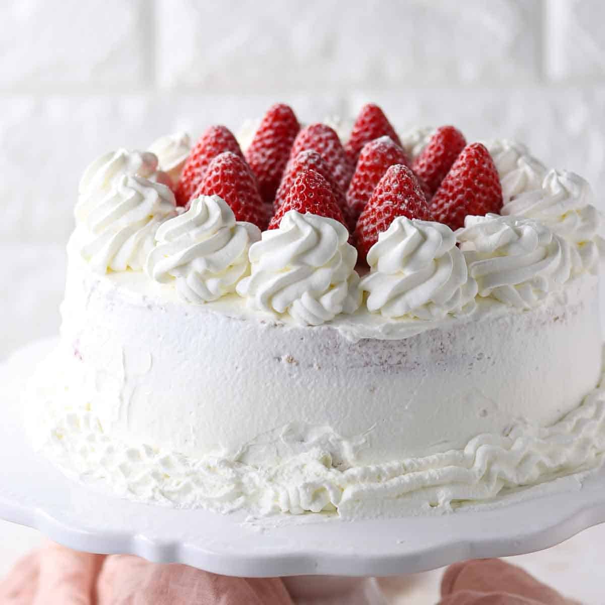 A round cake topped with whipped cream swirls and fresh strawberries on a white cake stand.