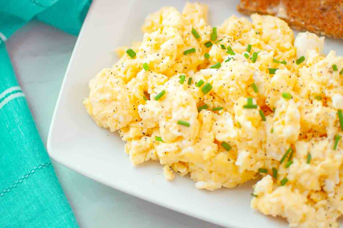 Scrambled eggs garnished with chopped chives and black pepper, served on a white plate next to a piece of toast.