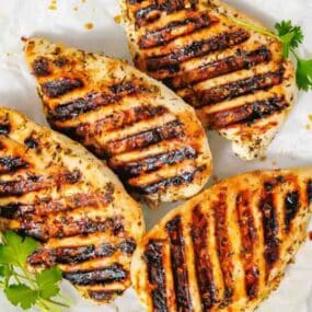 Four grilled chicken breasts with char marks on a white plate, garnished with sprigs of fresh cilantro.