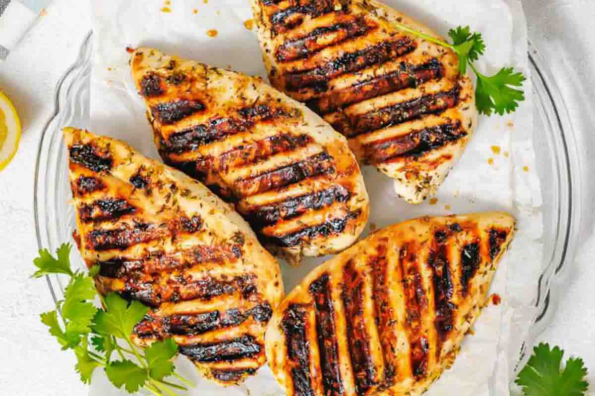 Four grilled chicken breasts with char marks on a white plate, garnished with sprigs of fresh cilantro.