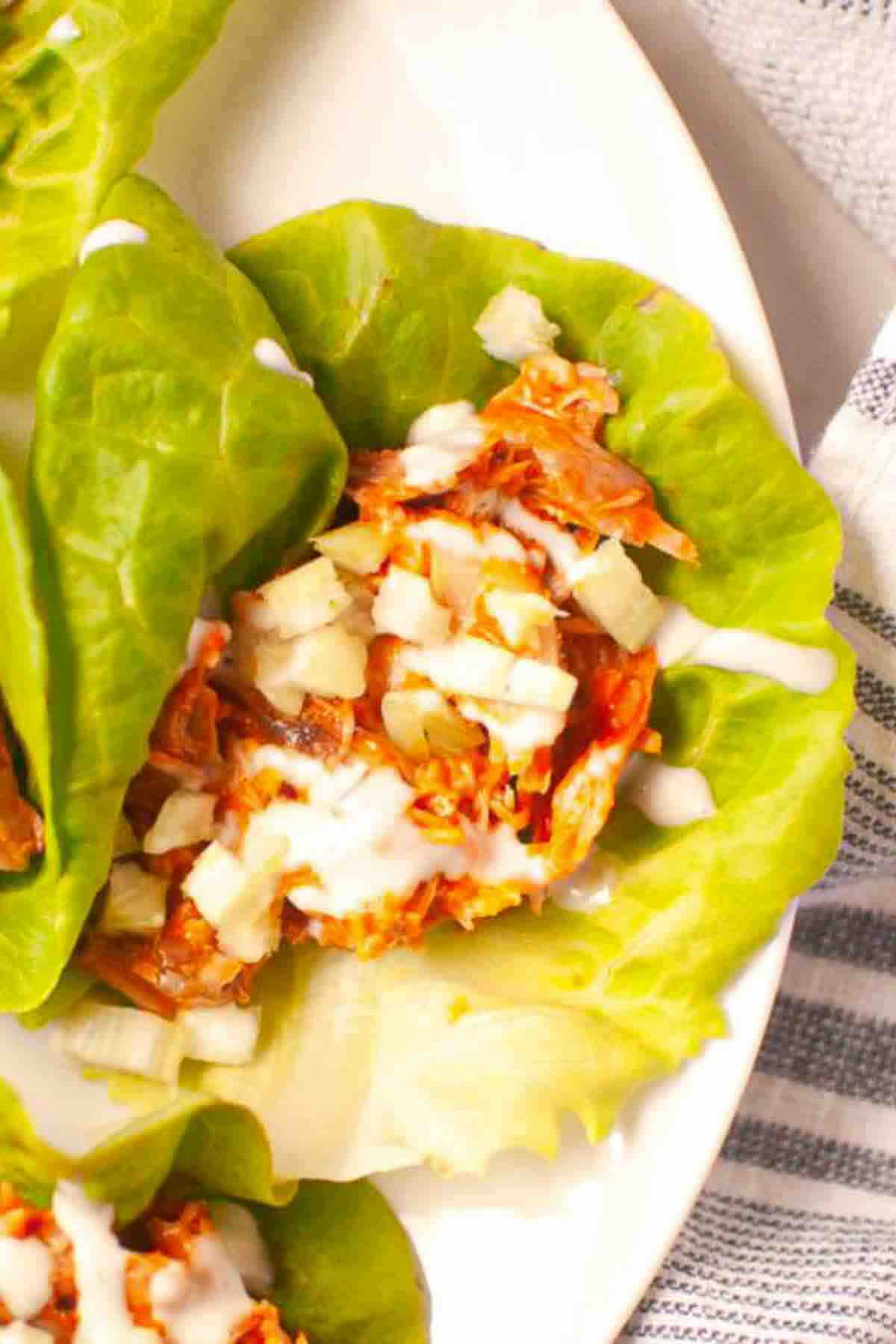 A close-up of a lettuce leaf filled with shredded meat, diced onions, and drizzled with a creamy white sauce on a white plate.