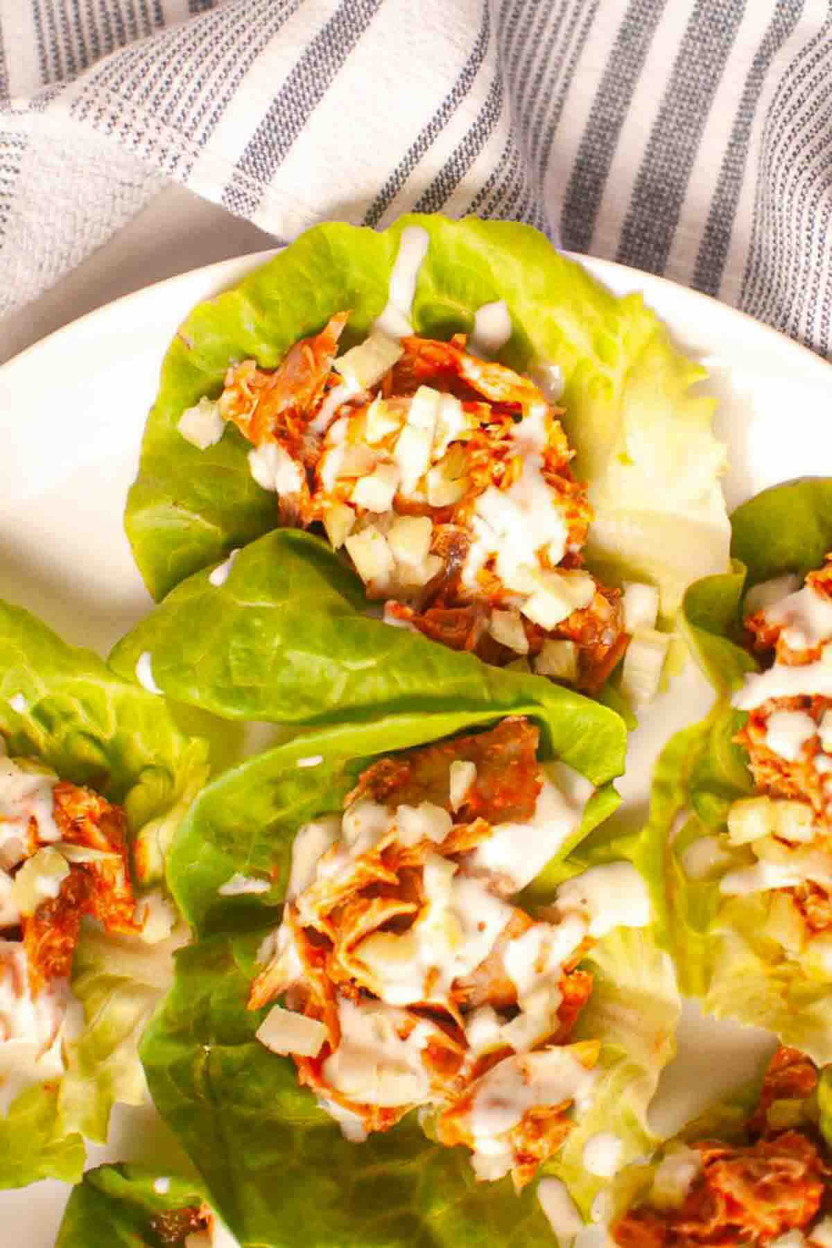Chicken lettuce wraps topped with chopped onions and drizzled with white sauce, served on a white plate next to a striped cloth.