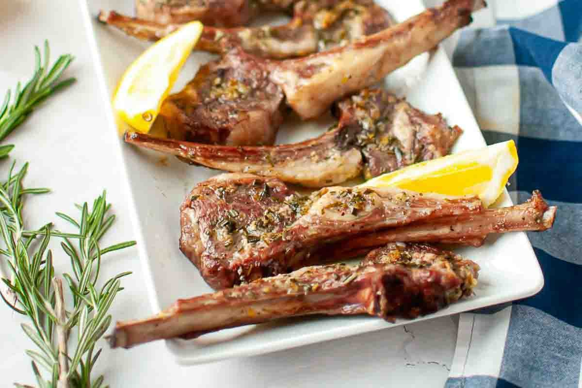 Grilled lamb chops garnished with herbs, served on a white rectangular plate with lemon wedges.