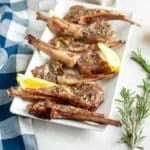 Rectangular white plate with grilled lamb chops, garnished with herbs and served with lemon wedges. A sprig of fresh rosemary is nearby on the table.