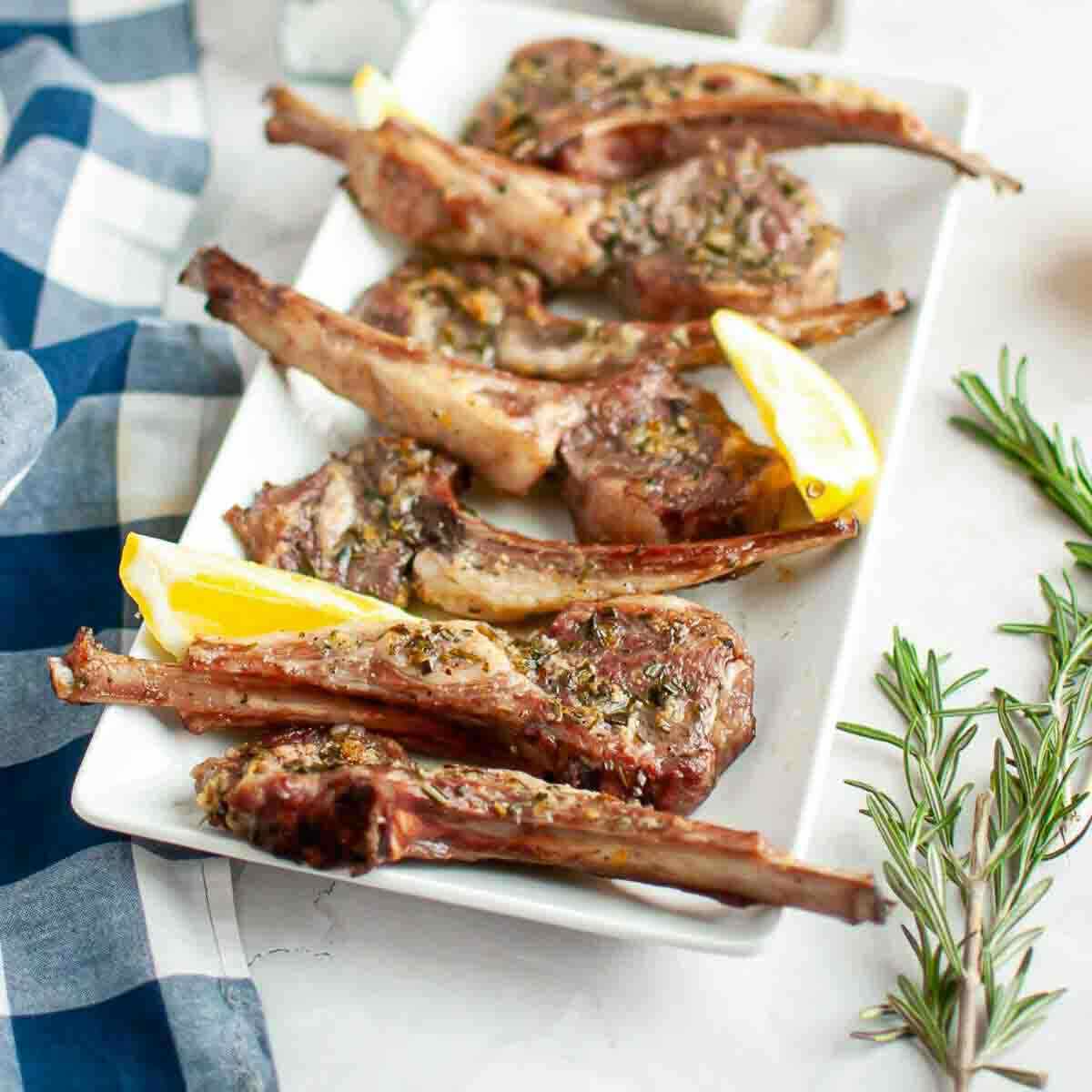 Rectangular white plate with grilled lamb chops, garnished with herbs and served with lemon wedges. A sprig of fresh rosemary is nearby on the table.