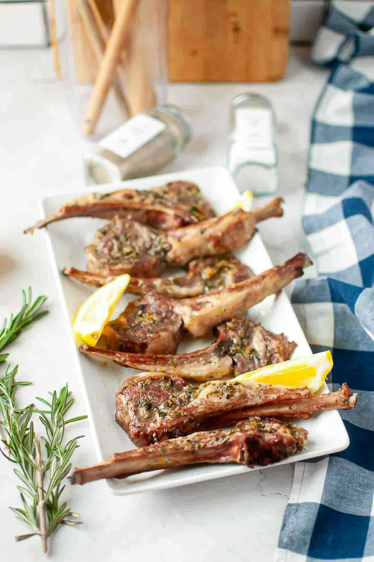 A rectangular white plate with six grilled lamb chops, garnished with herbs and lemon wedges, sits on a light countertop next to rosemary, spice jars, and a blue checkered cloth.
