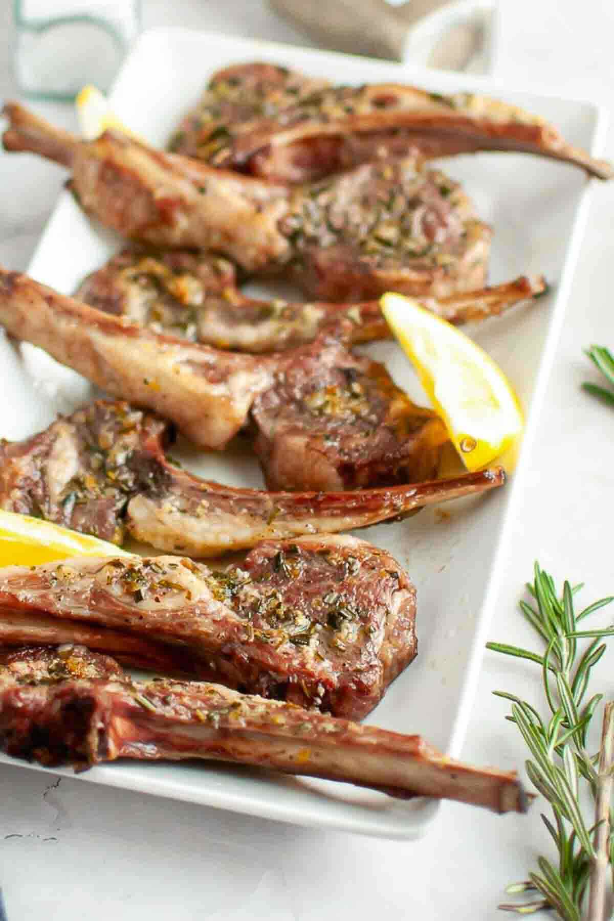 Grilled lamb chops garnished with herbs and lemon wedges are arranged on a rectangular white plate.