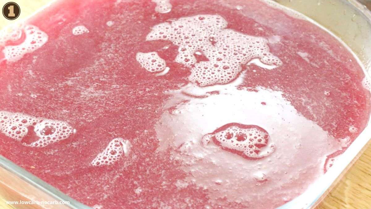 A close-up view of a glass dish filled with a red liquid mixture with bubbles and foam on the surface.