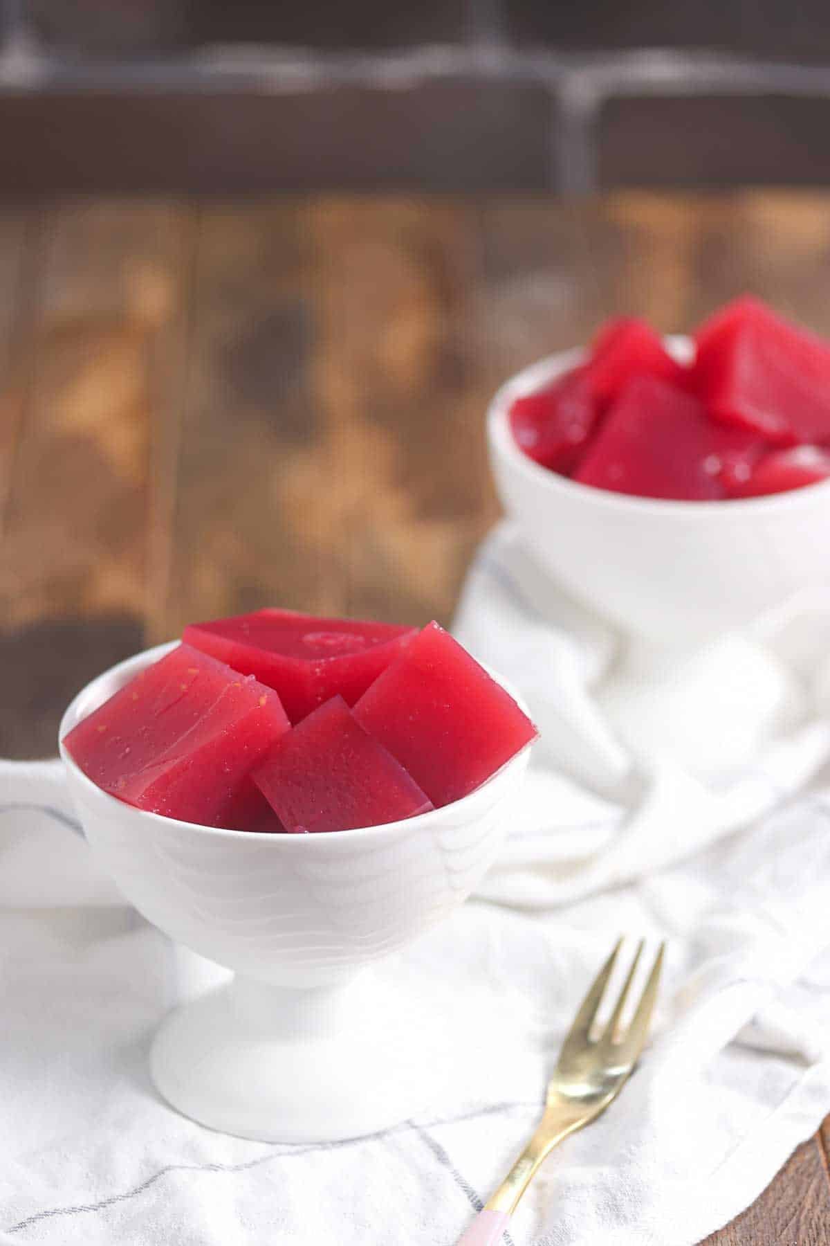 Two white dessert bowls filled with red gelatin cubes are placed on a white cloth, with a gold fork nearby on a wooden surface.