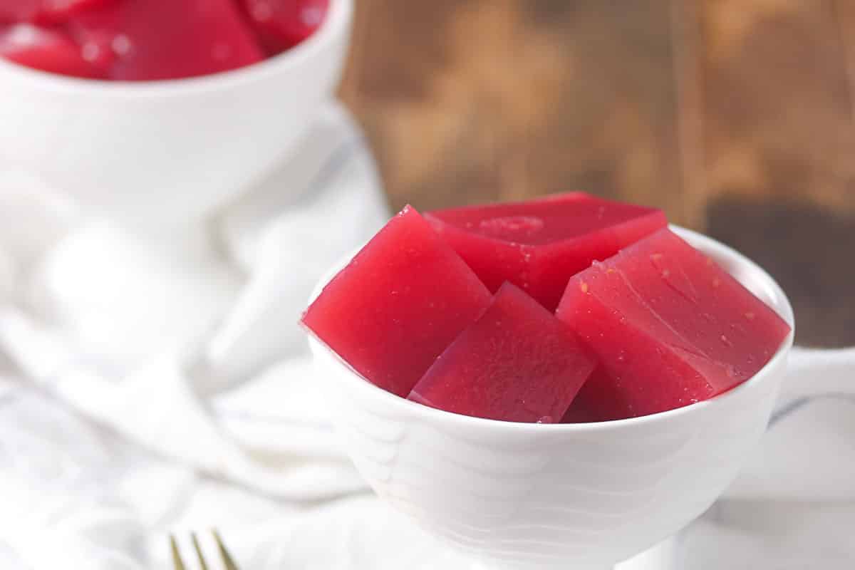 A white bowl filled with red gelatin cubes sits on a white cloth, with another bowl of gelatin in the background.