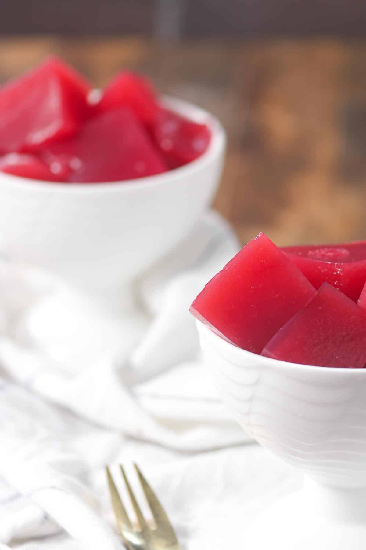 Two white bowls filled with red, translucent gelatin cut into square pieces, placed on a white cloth with a fork nearby.