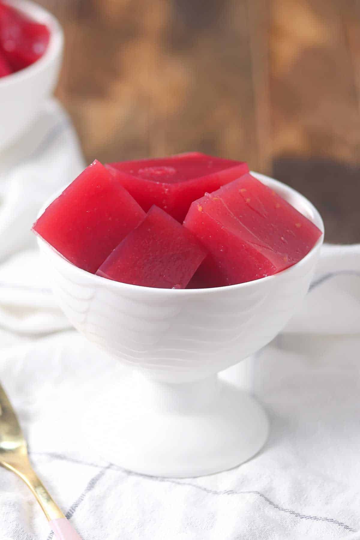 A white bowl filled with red gelatin cubes, placed on a white cloth with a gold spoon beside it.