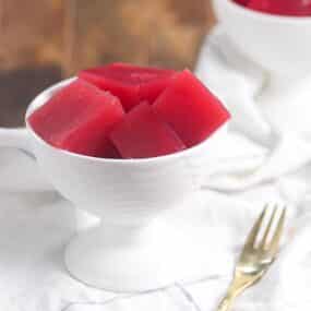 A white dessert bowl filled with red gelatin cubes sits on a white cloth next to a gold fork.