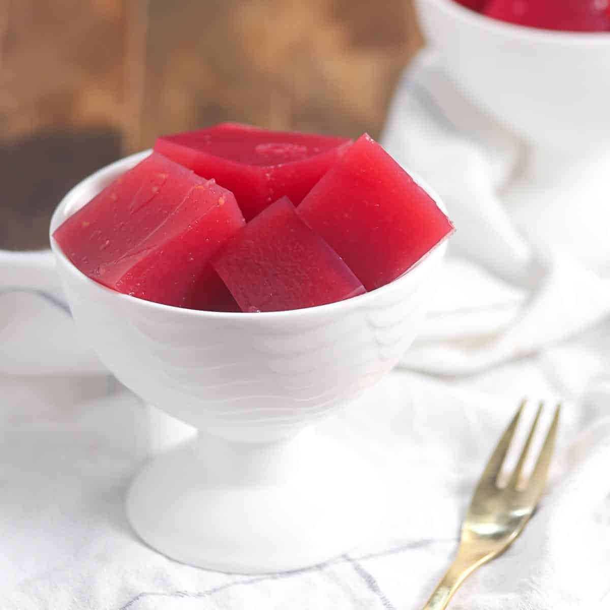 A white dessert bowl filled with red gelatin cubes sits on a white cloth next to a gold fork.