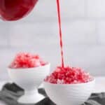 Red syrup being poured onto shaved ice in a white bowl, with another bowl of shaved ice in the background.