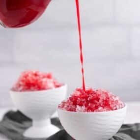 Red syrup being poured onto shaved ice in a white bowl, with another bowl of shaved ice in the background.