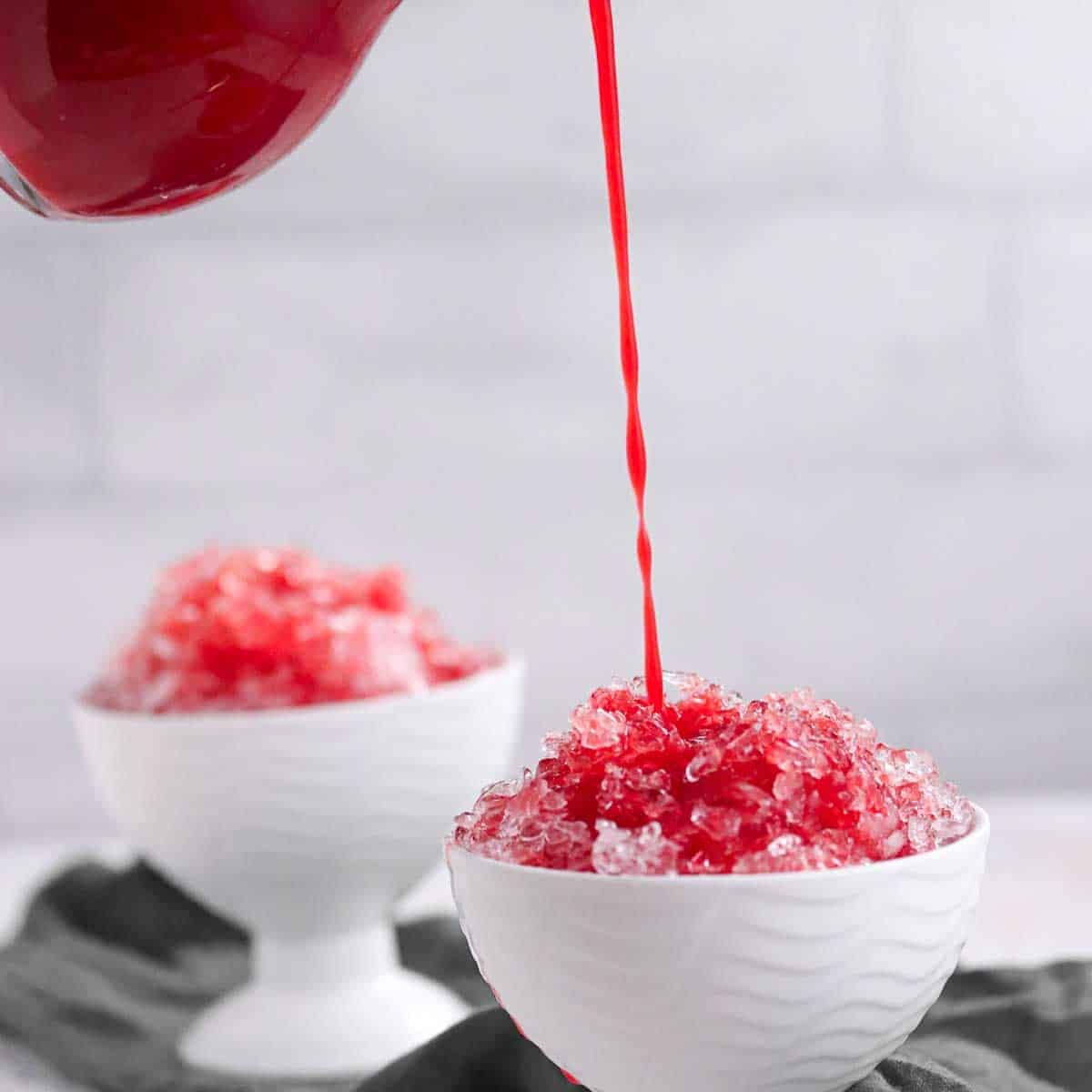 Red syrup being poured onto shaved ice in a white bowl, with another bowl of shaved ice in the background.