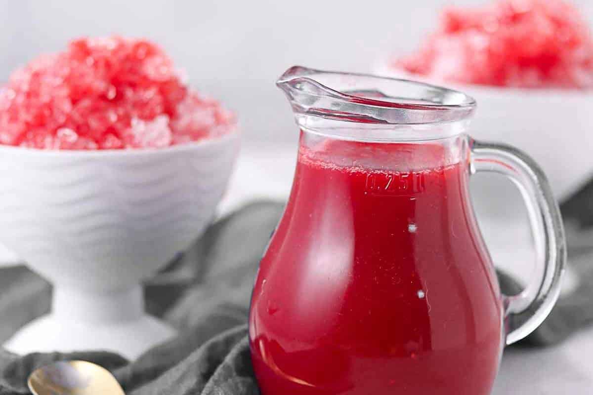 A glass pitcher filled with red syrup sits on a gray cloth, with a white bowl of red shaved ice in the background.
