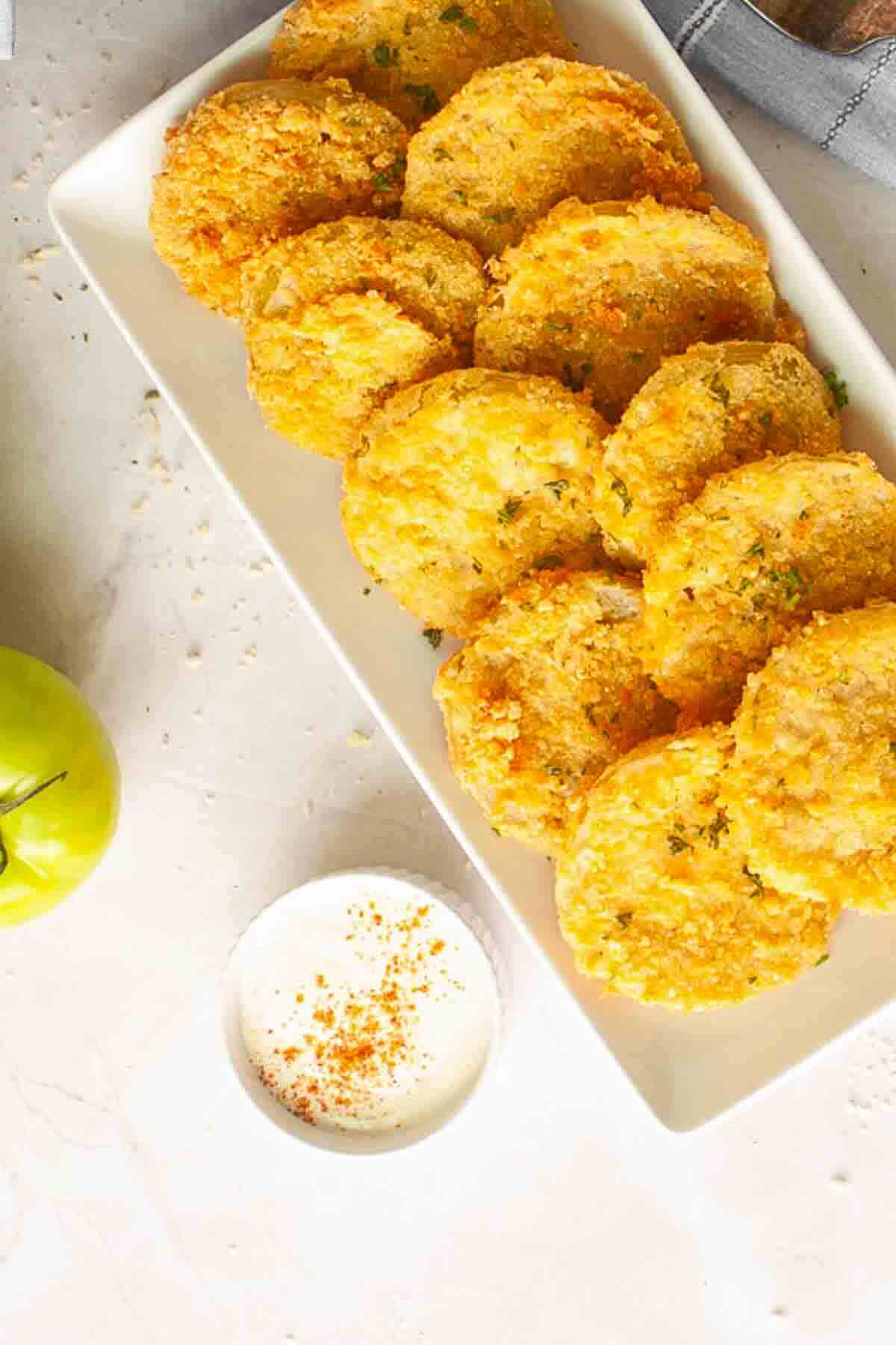 A rectangular white plate with several golden fried green tomato slices, next to a small bowl of dipping sauce and a whole green tomato on a light surface.