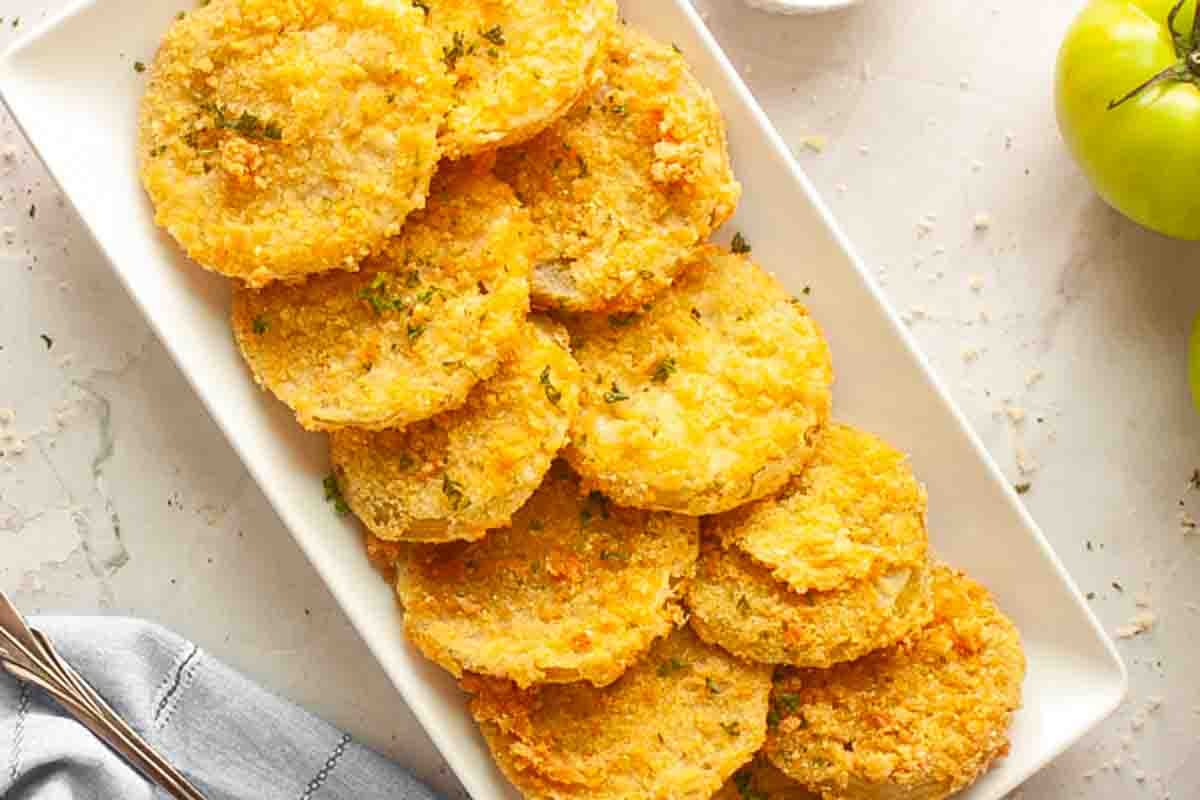 A rectangular white plate holds a neatly arranged stack of golden-fried green tomato slices, garnished with herbs.