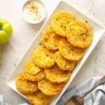 A rectangular white plate with a neatly arranged stack of golden-fried green tomato slices, garnished with herbs, next to a small bowl of dipping sauce.