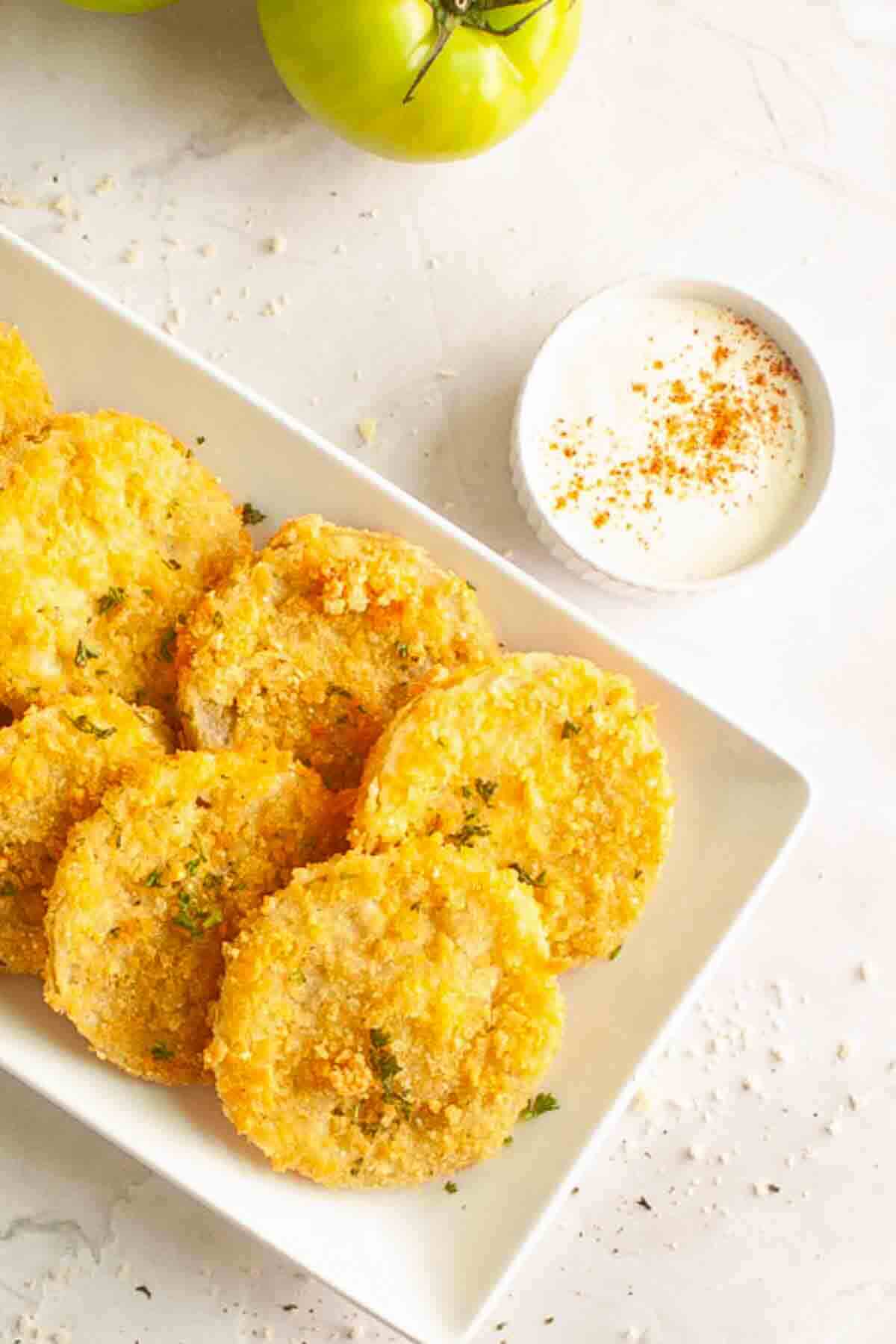 A plate of golden, breaded fried green tomatoes is served with a small bowl of creamy dipping sauce on a white surface.