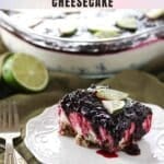 A slice of lemon and blackberry cheesecake topped with lime slices is served on a white plate; the whole cheesecake is in the background.