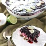 A square slice of berry dessert with a crumb crust, creamy middle layer, and berry topping, garnished with lime wedges, sits on a white plate next to a fork and knife.