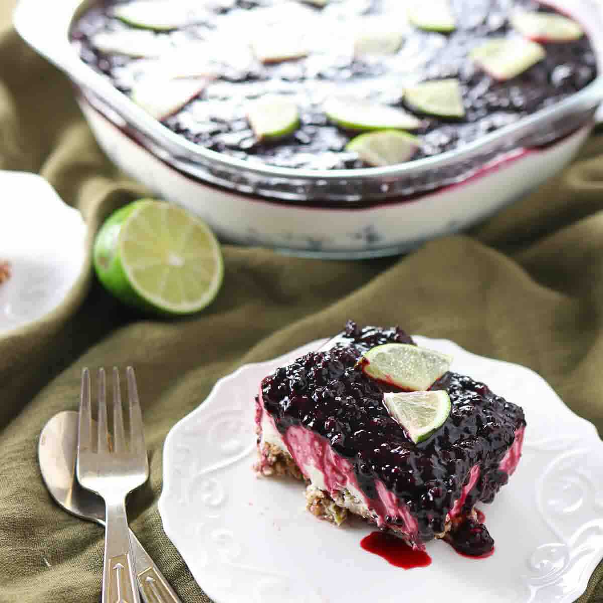 A square slice of berry dessert with a crumb crust, creamy middle layer, and berry topping, garnished with lime wedges, sits on a white plate next to a fork and knife.