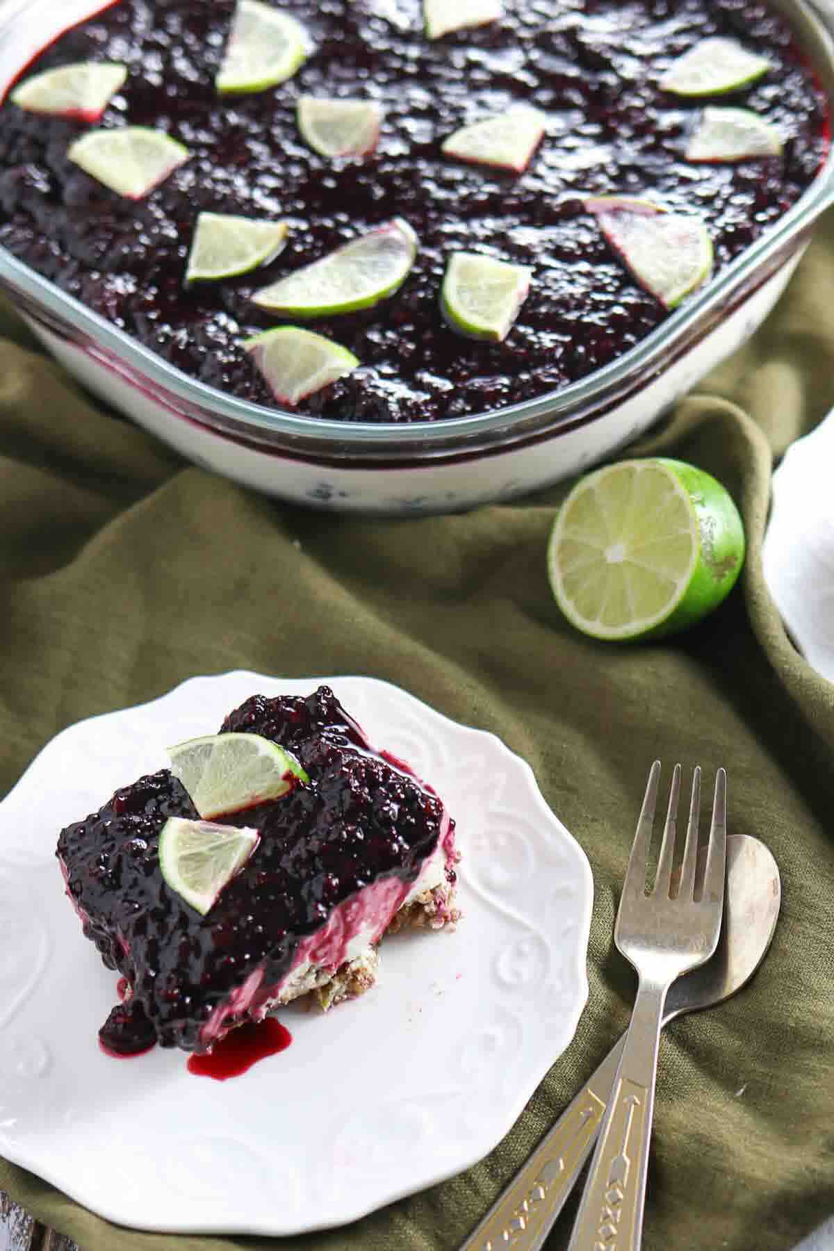 A slice of layered dessert with a dark berry topping and lime wedges on a white plate, with the rest of the dessert in a glass dish nearby. Fork and spoon placed beside the plate.