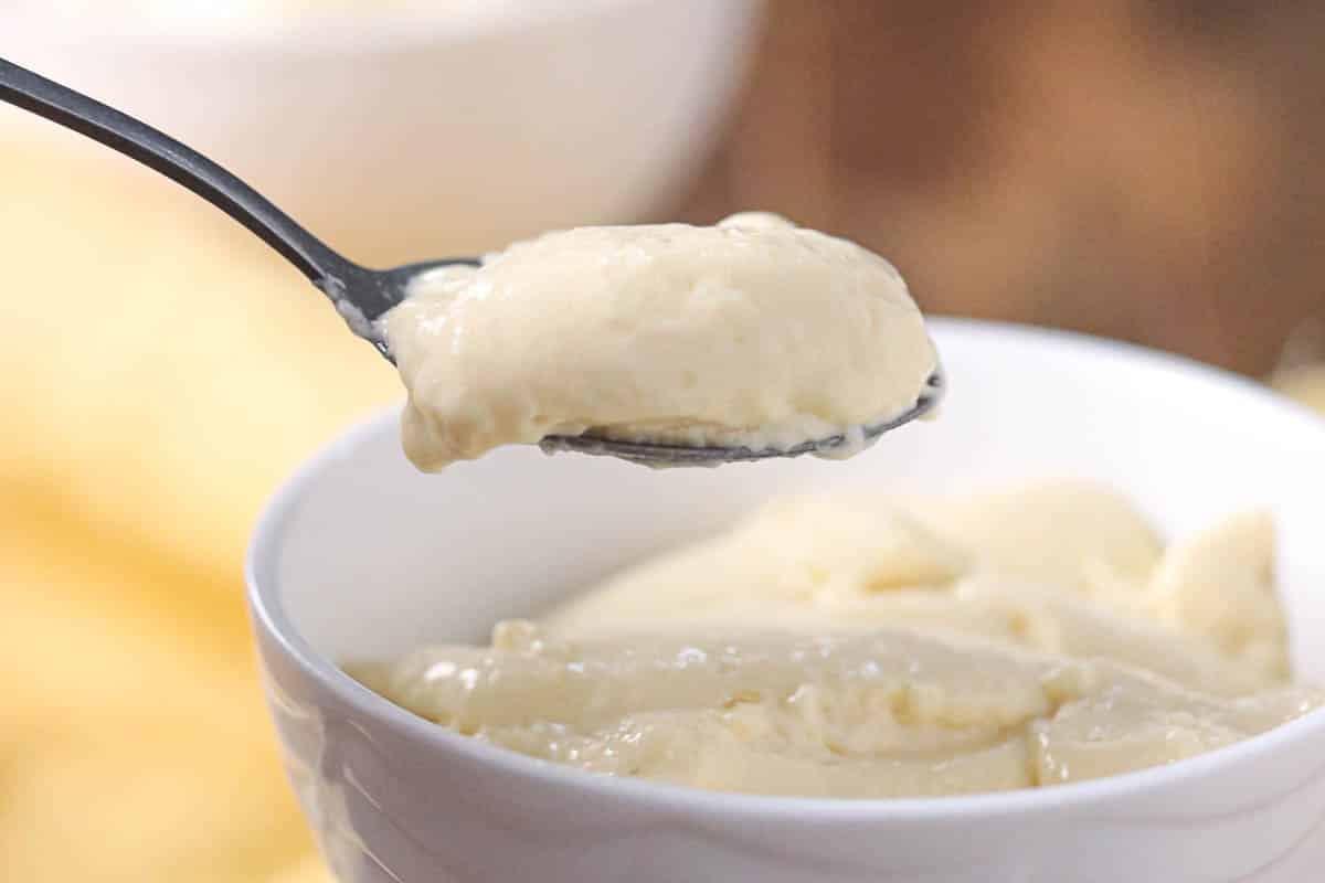 A spoonful of creamy banana pudding held above a white bowl filled with more pudding, with a blurred background.