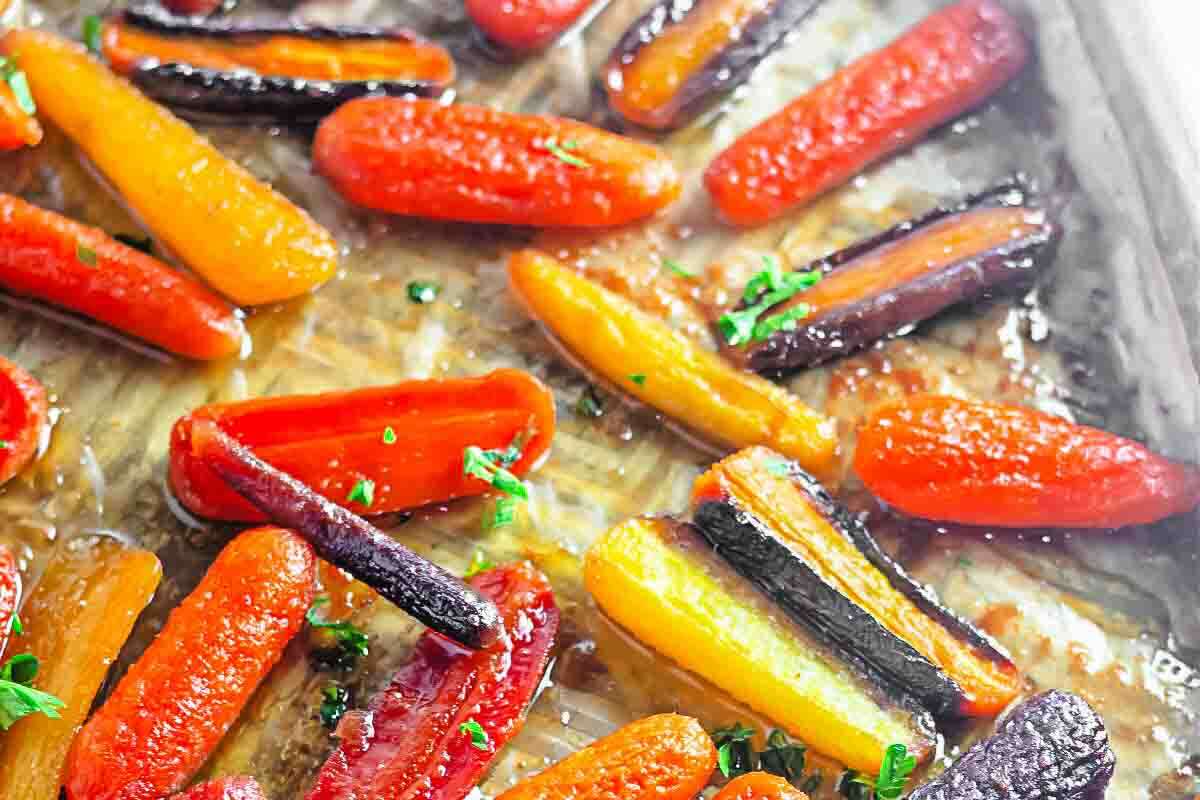 Assorted roasted carrots in orange, yellow, and purple colors arranged on a baking sheet with a light glaze and sprinkled with chopped herbs.