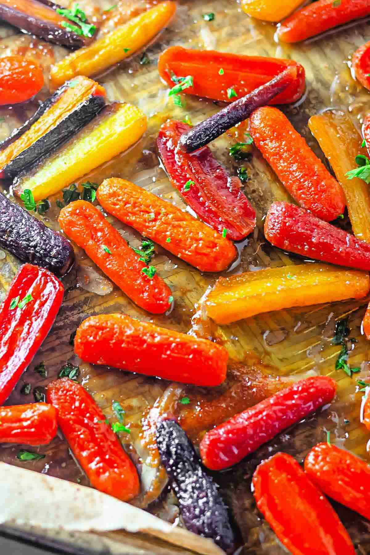 Roasted baby carrots in orange, purple, and yellow varieties on a baking sheet, garnished with chopped herbs and a drizzle of glaze.