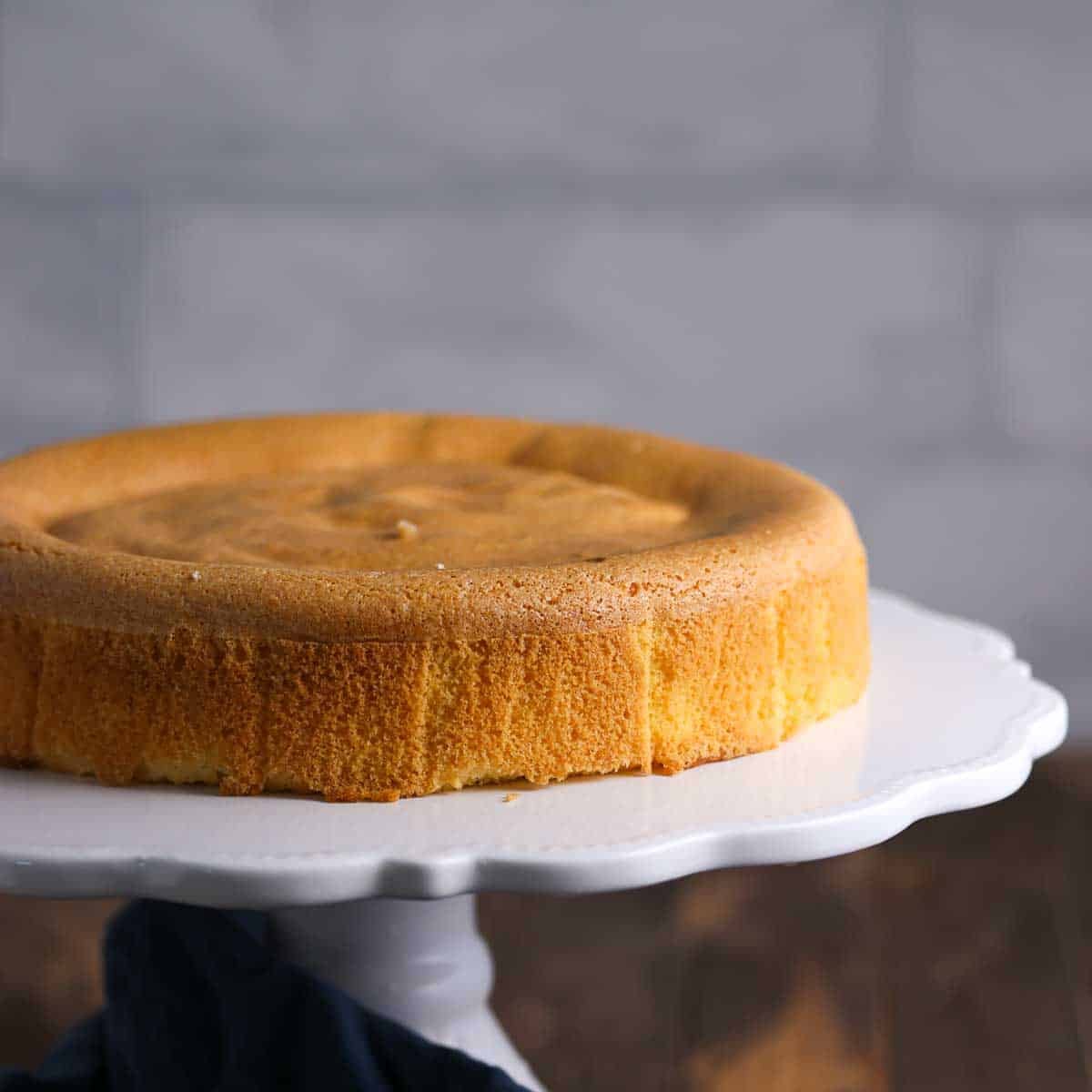 A plain, golden-brown sponge cake sits on a white cake stand against a blurred stone background.