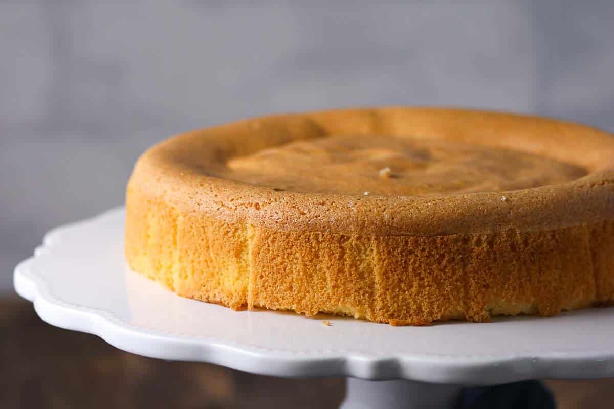 A golden brown sponge cake with a slightly sunken center sits on a white cake stand against a neutral background.