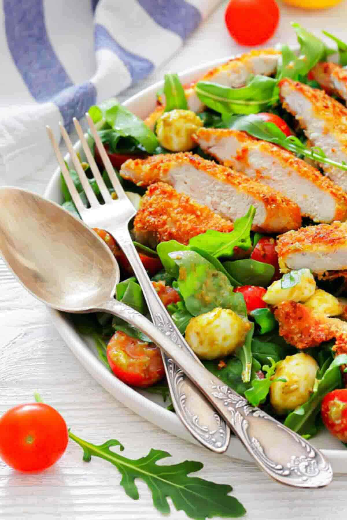 A plate of salad with breaded chicken slices, cherry tomatoes, mozzarella balls, and arugula, next to a fork and spoon on a white table.