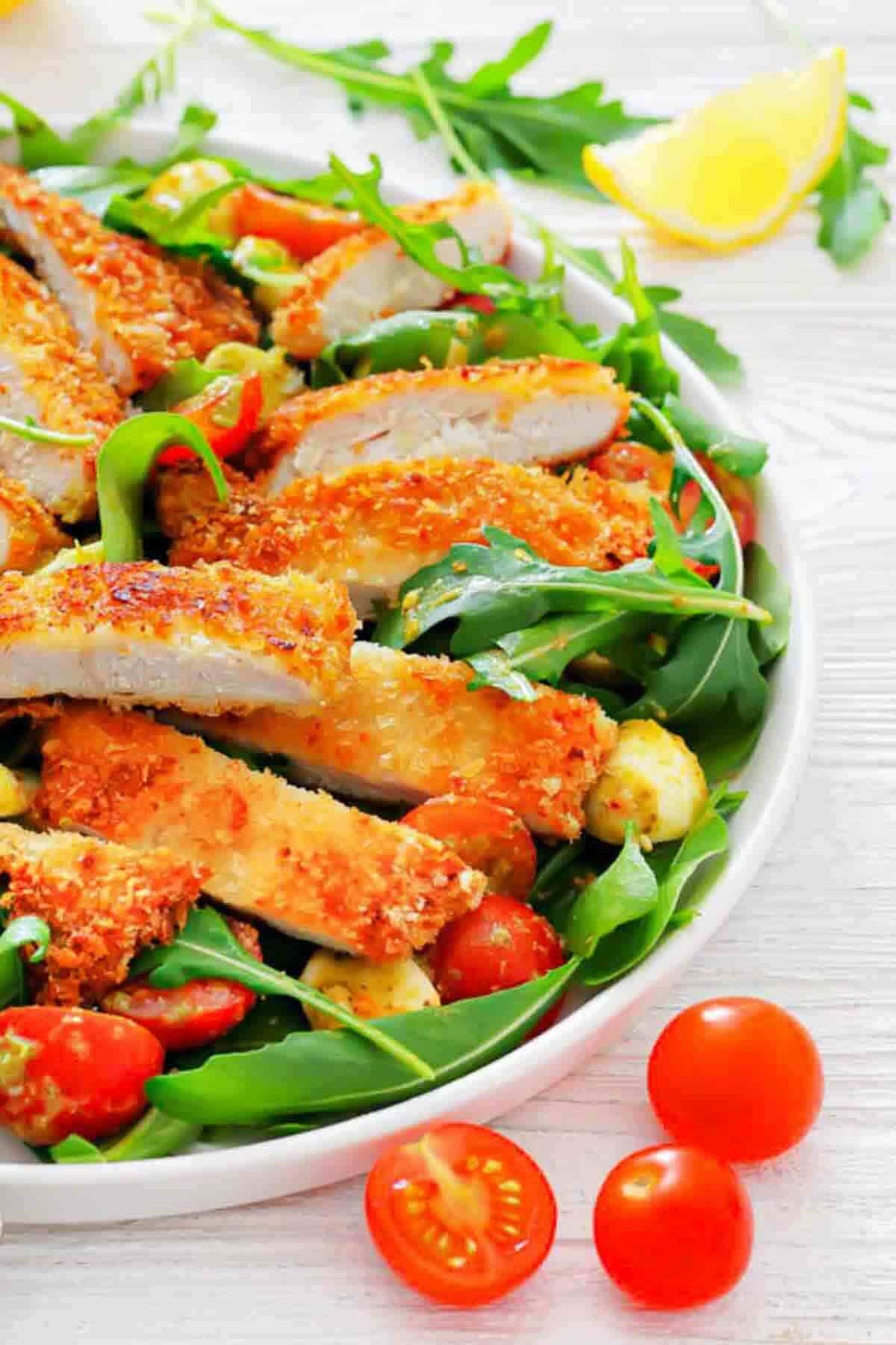 Sliced breaded chicken served on a bed of arugula with cherry tomatoes and mozzarella balls, garnished with lemon wedges.