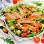A plate of breaded chicken slices on a bed of arugula, cherry tomatoes, and mozzarella balls, with serving utensils on the side.