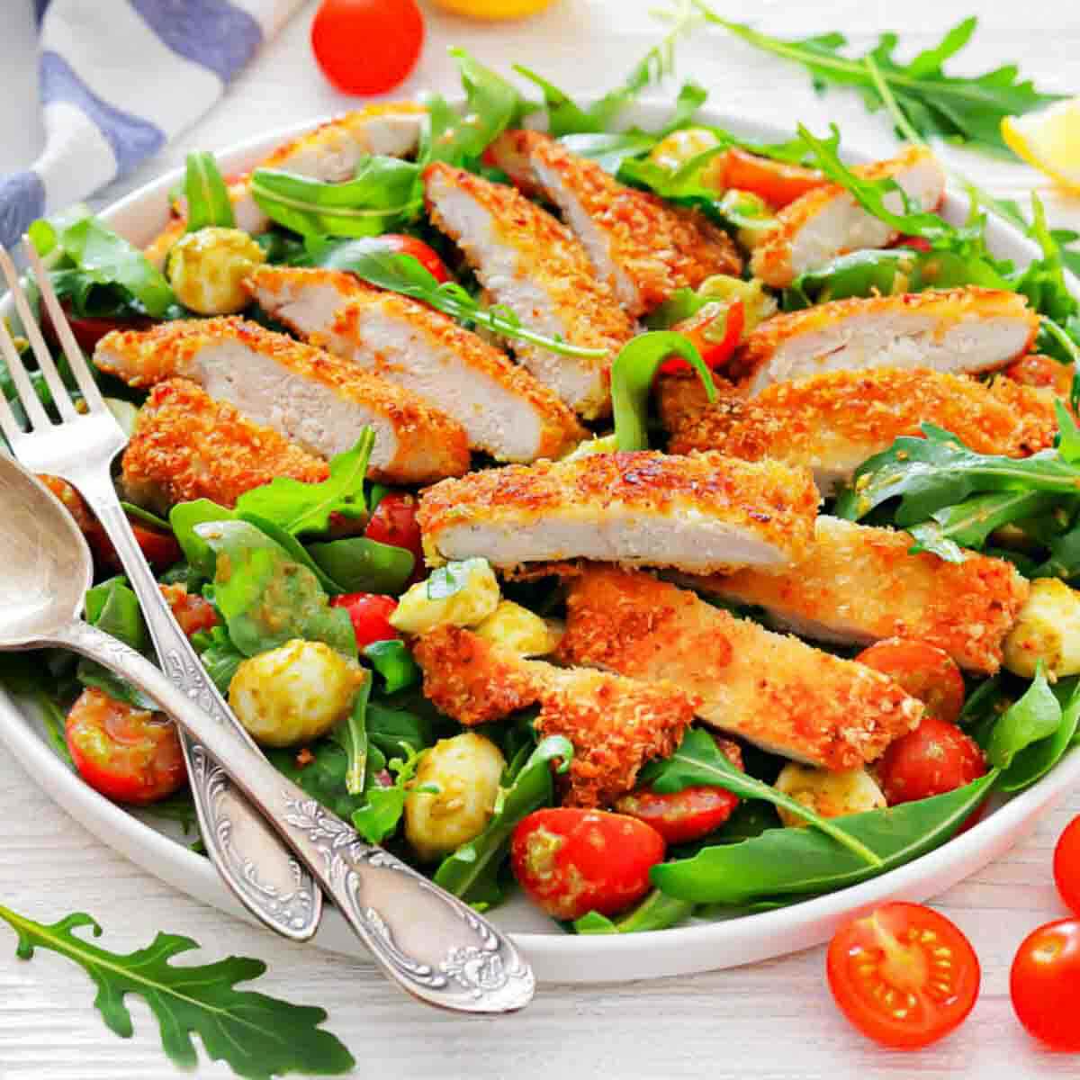 A plate of breaded chicken slices on a bed of arugula, cherry tomatoes, and mozzarella balls, with serving utensils on the side.