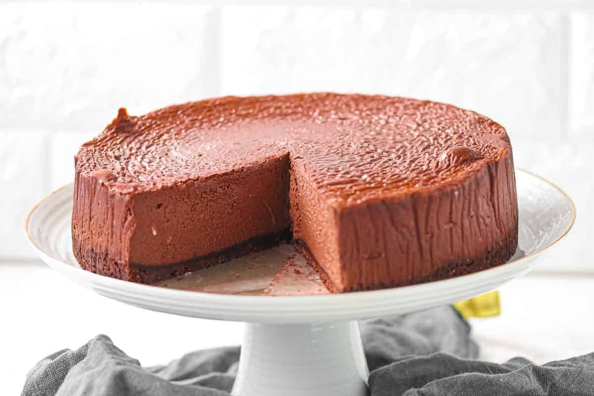 Chocolate cheesecake with a smooth top sits on a cake stand, with one slice removed to reveal the creamy interior.