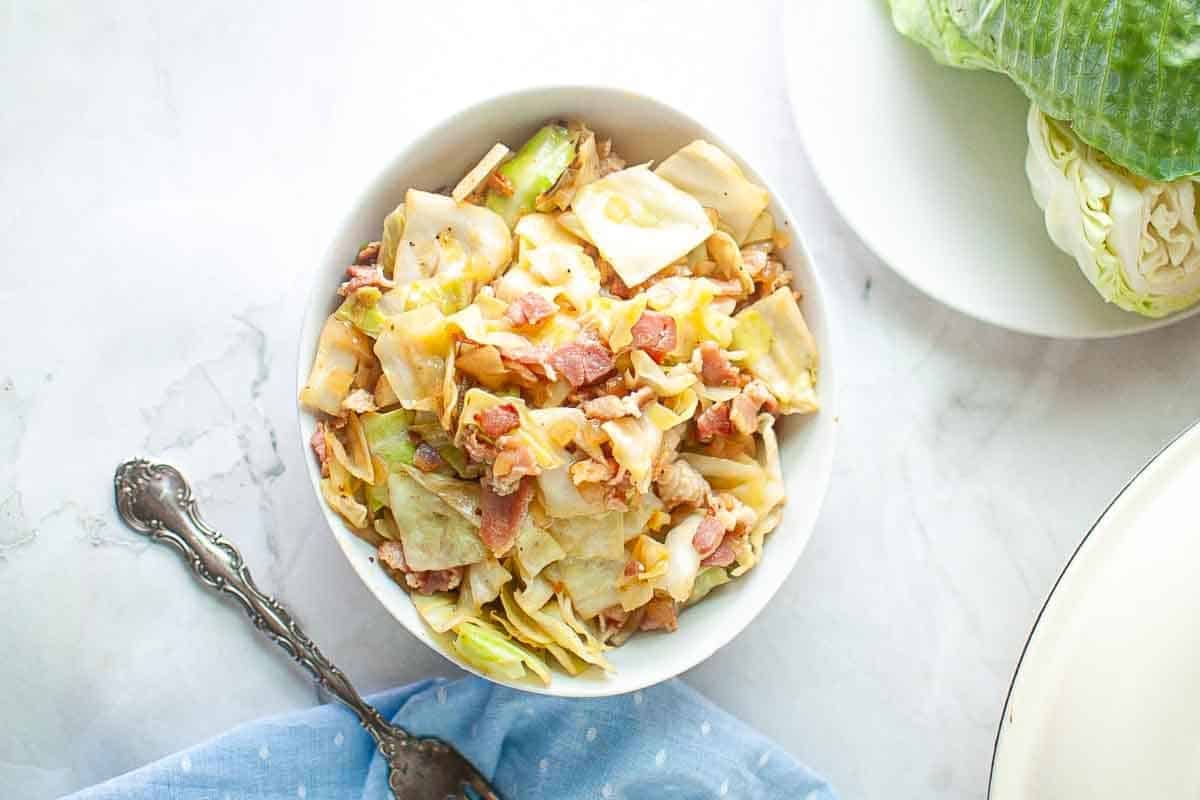 A white bowl filled with chopped cooked cabbage and bacon pieces sits on a marble surface next to a vintage fork and a partially visible cabbage head.