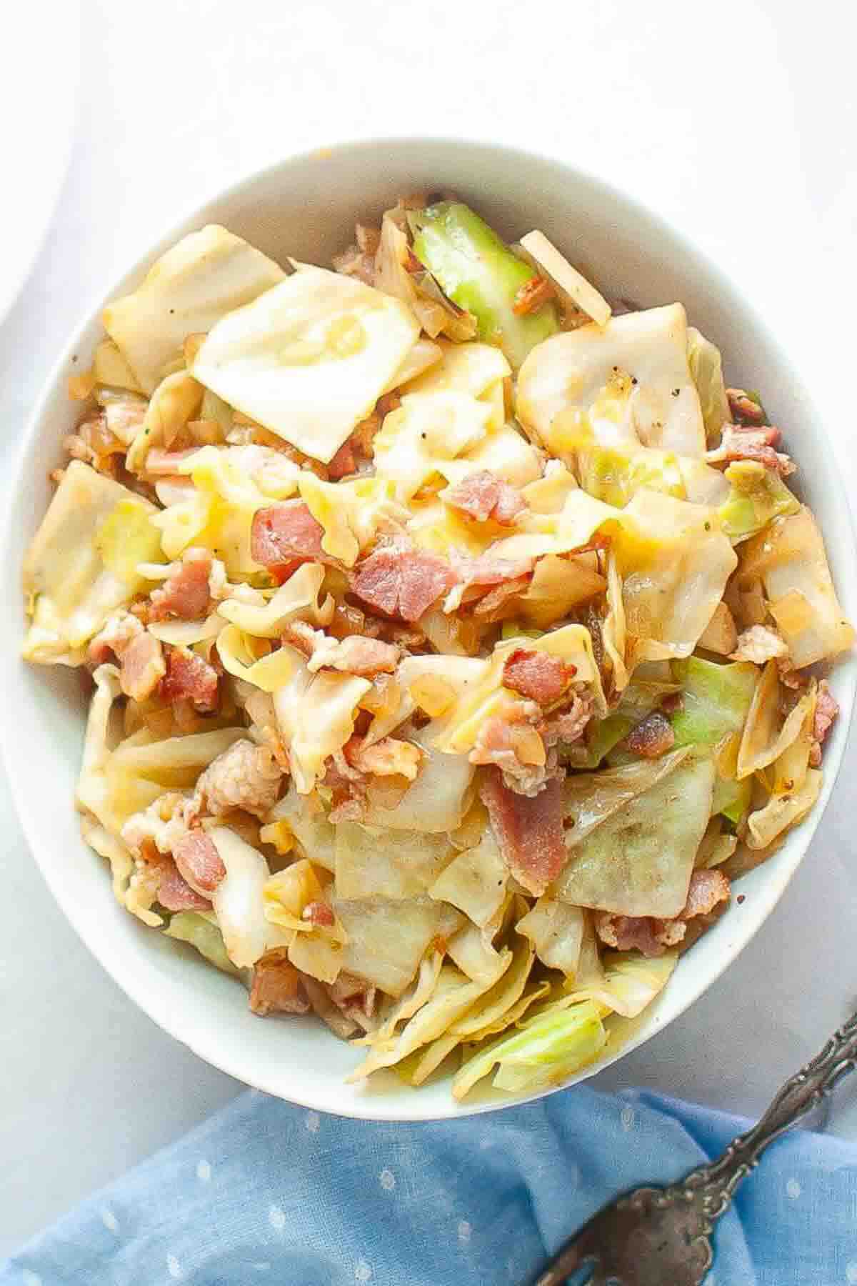 A bowl filled with cooked cabbage and bacon pieces, placed on a white surface with a blue napkin and a spoon nearby.