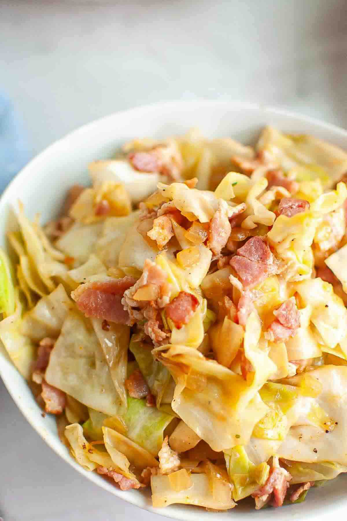 A bowl of cooked cabbage mixed with pieces of bacon, served in a white dish on a light surface.