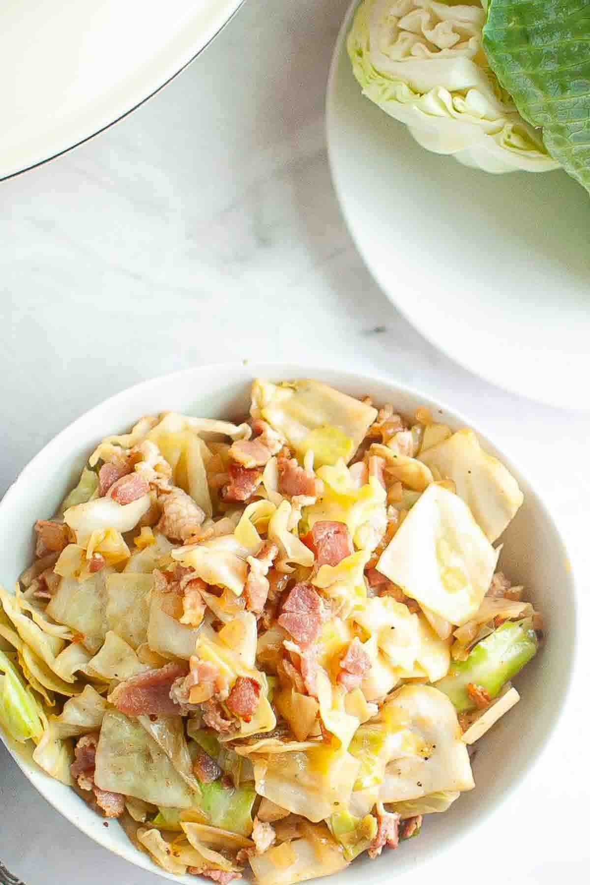 A bowl of cooked cabbage mixed with pieces of bacon sits on a white surface next to a plate with a partially cut cabbage.