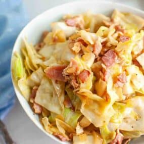 A bowl of cooked cabbage mixed with pieces of bacon, seasoned and served in a white dish.