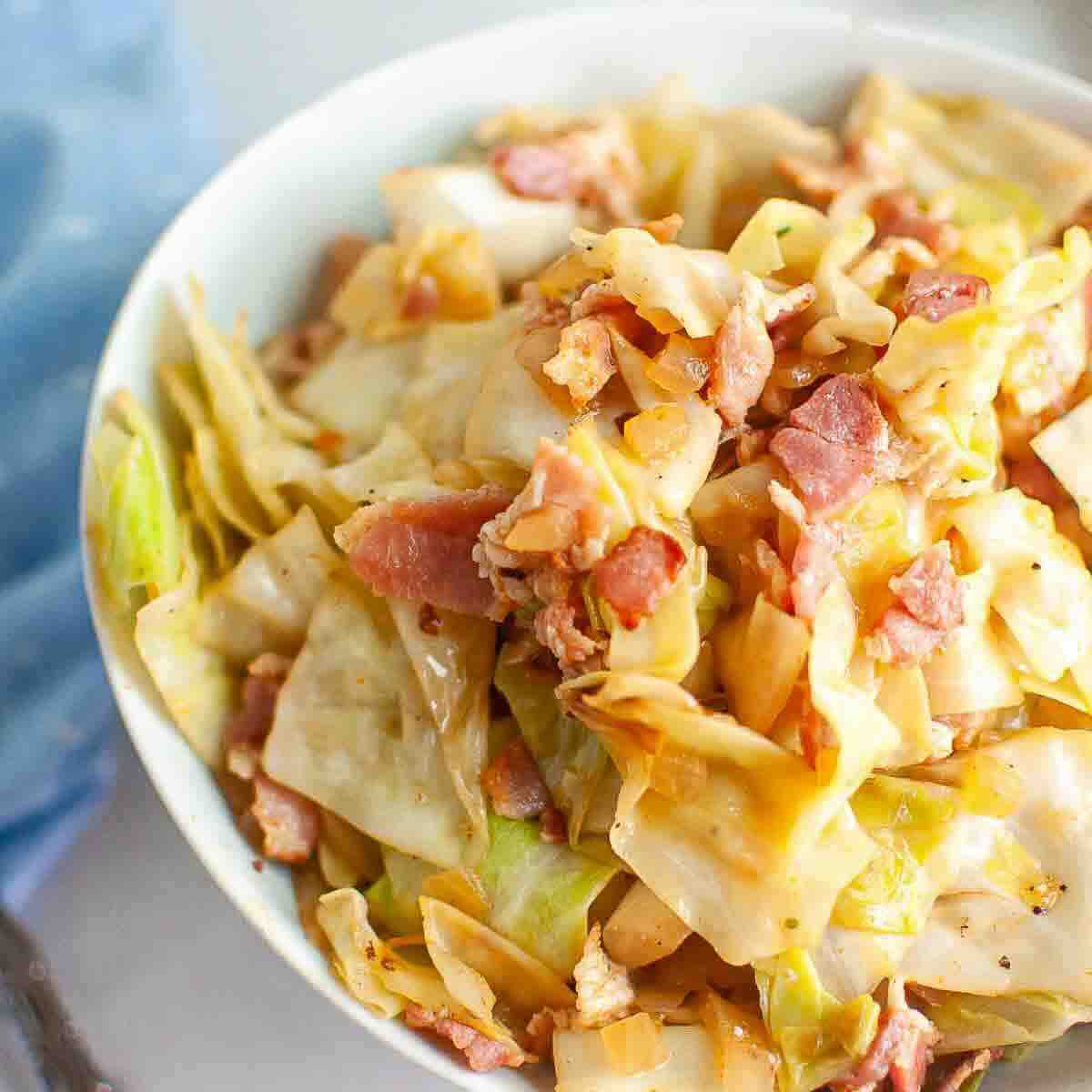 A bowl of cooked cabbage mixed with pieces of bacon, seasoned and served in a white dish.