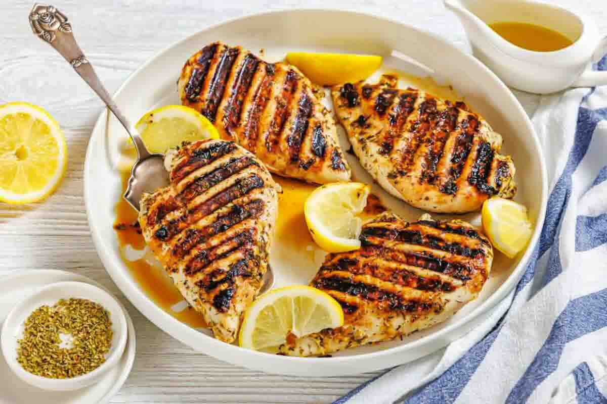 Four grilled chicken breasts are served on a round plate with lemon wedges, a spoon, seasoning, and a small jug of sauce on the side.