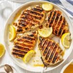 Grilled chicken breasts with visible char marks served on a white plate, garnished with lemon wedges.