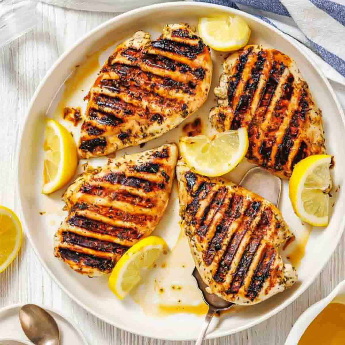 Grilled chicken breasts with visible char marks served on a white plate, garnished with lemon wedges.
