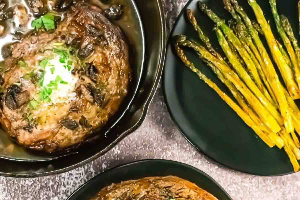 Cooked steak with mushrooms and herbs in a skillet, served alongside a plate of roasted asparagus spears on a dark surface.